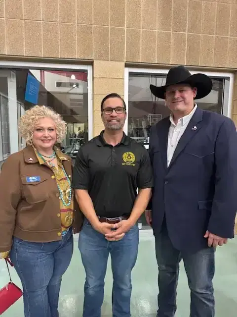 A woman in a brown jacket poses indoors with two men, one wearing a black cowboy hat.