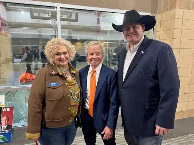 A woman in a brown jacket poses with two men, one in an orange tie, one in a cowboy hat.