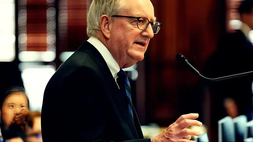 John Smithee, a gray-haired man in glasses and a dark suit, speaking at a microphone in a legislative chamber.