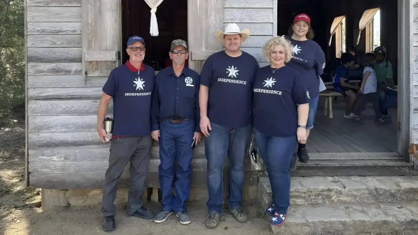 Five TNM members in matching 'Independence' shirts stand in front of a weathered wooden hall at Washington-on-the-Brazos.