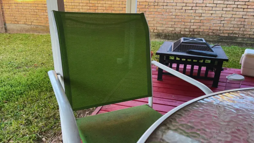 Empty green mesh patio chair beside a glass table on a red deck, with a fire pit behind it.