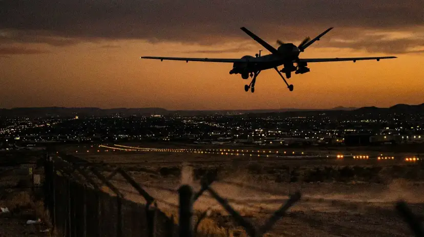 A large military drone silhouetted against an orange sunset as it flies low over a lit airport runway.