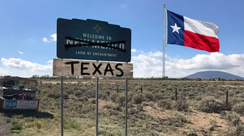 A 'Welcome to New Mexico' sign with New Mexico crossed out and a 'Texas' board below, beside a Texas flag.