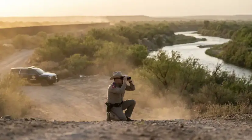 A Texas trooper in a cowboy hat kneels with binoculars on a dusty rise above a river, a patrol SUV behind him.