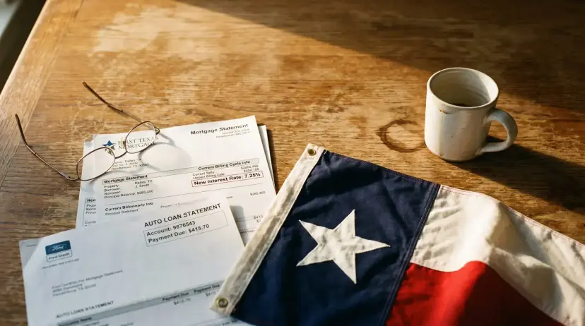 Mortgage and auto loan statements lie beside reading glasses and a coffee mug, with a Texas flag on the wooden table.