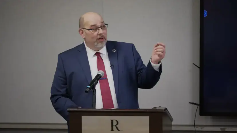 A bald man with glasses in a blue suit and red tie speaking and gesturing at a podium.