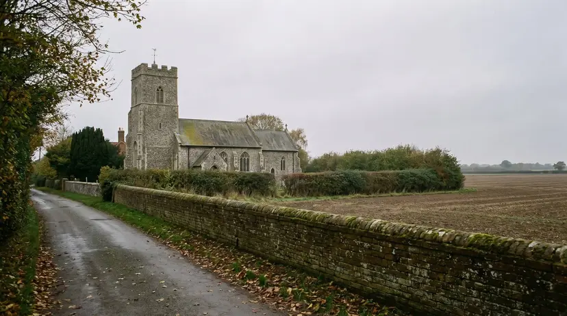 An old flint parish church with a square tower beside a narrow lane and brick wall in rural Suffolk on an overcast autumn day.