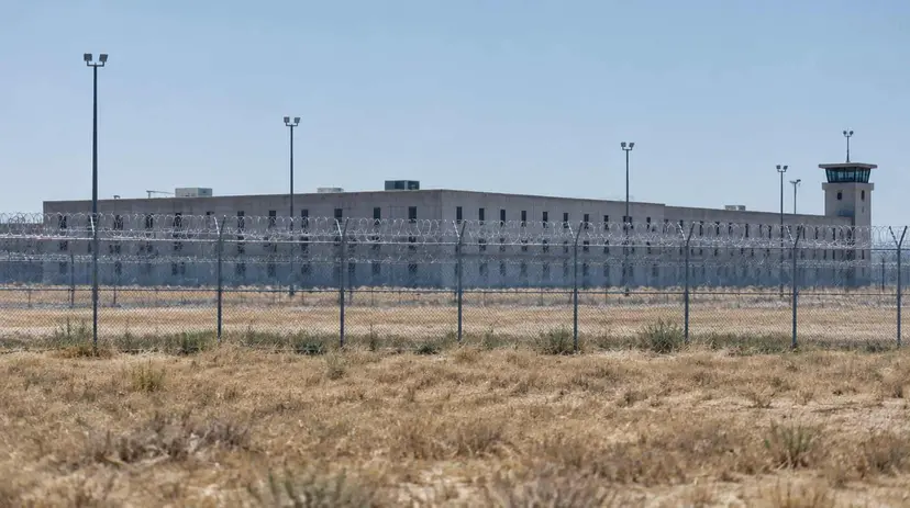A low concrete prison building behind a chain-link fence topped with razor wire under a hot, hazy sky.