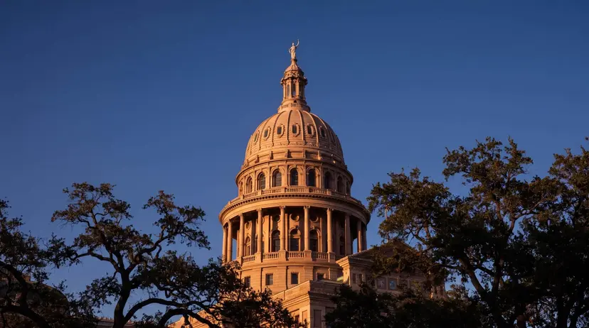 The dome of the Texas State Capitol in Austin, topped by the Goddess of Liberty, lit at sunset between live oaks.
