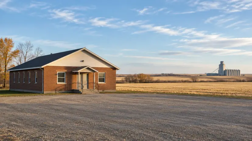 A brick community hall beside harvested wheat fields and a grain elevator in a small Alberta prairie town.