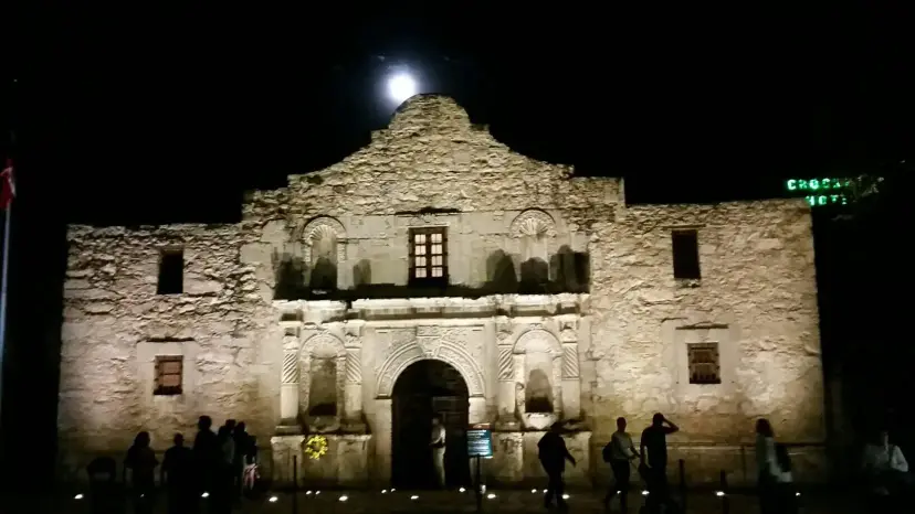 The Alamo church lit up at night under a full moon, with visitors silhouetted in front.