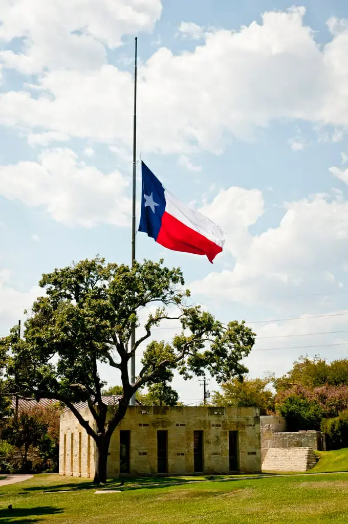 Texas flag flying at half-staff over a limestone building and a large oak tree.