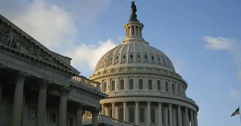 The U.S. Capitol dome in Washington beside a columned wing of the building under a blue sky.
