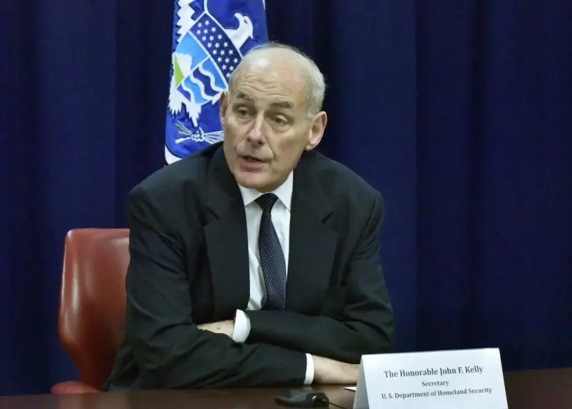 Federal Homeland Security Secretary John F. Kelly seated at a table behind his nameplate, with a flag behind him.