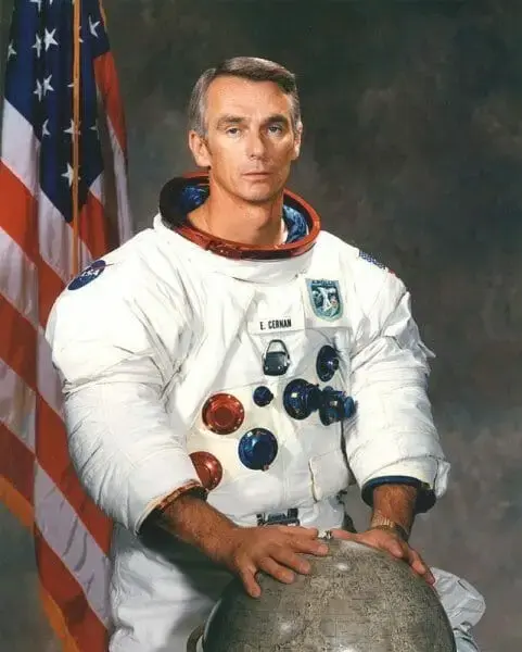 Astronaut Gene Cernan in a white spacesuit, one hand on a lunar globe, beside an American flag.