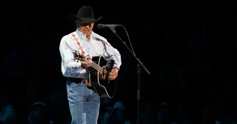 George Strait in a black cowboy hat playing an acoustic guitar at a microphone on a dark stage.