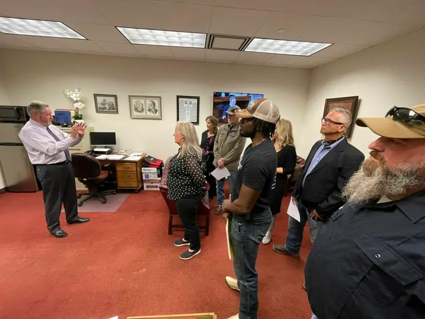 A man in a white shirt and tie speaks to a group of visitors standing in a Capitol office.