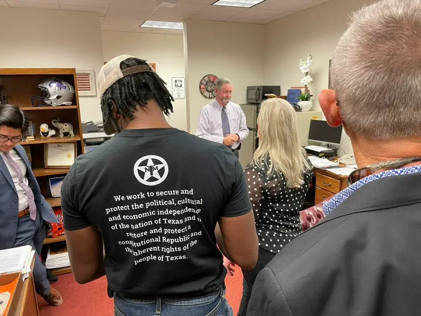 A man in a Texas Nationalist Movement mission statement shirt listens to a legislator in his office.