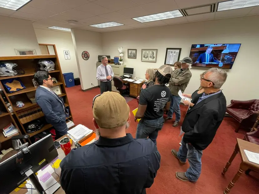 Visitors wearing TNM shirts meet with a legislator in an office with a TV showing the House floor.