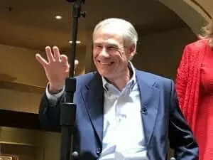 A smiling gray-haired man in a blue blazer waving from a stage beside a microphone stand.