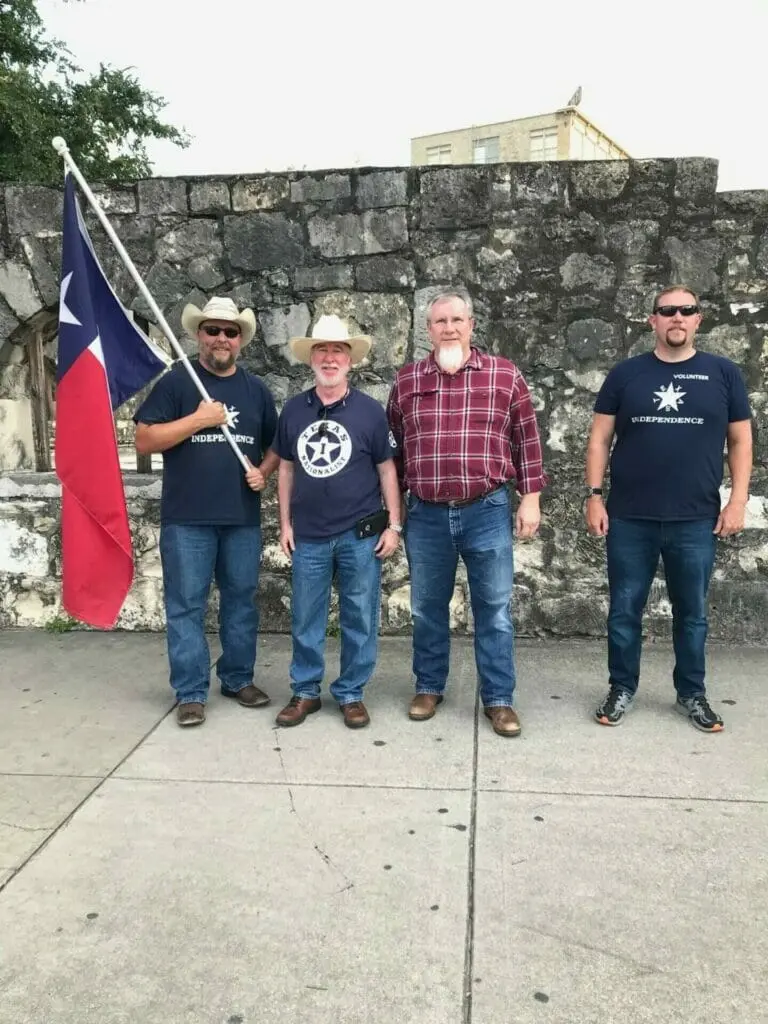 Four men, one holding a Texas flag, stand together in front of a limestone wall.