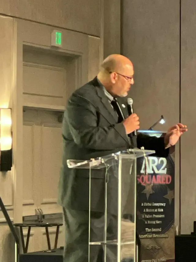 A man speaks at a clear podium with an 'AR2 Squared' banner in a hotel ballroom.