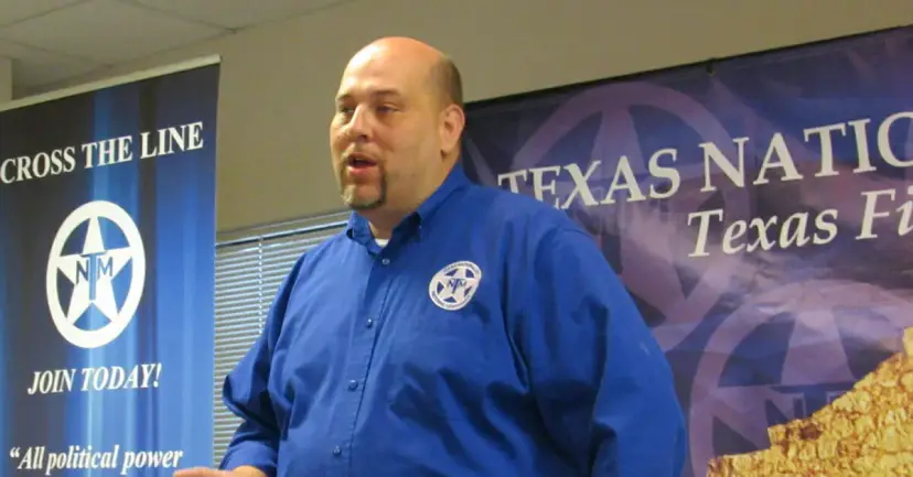 TNM President Daniel Miller speaking in a blue TNM shirt in front of Texas Nationalist Movement banners.