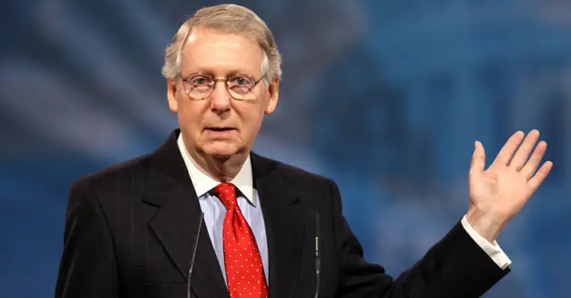 A gray-haired man in glasses and a red tie raising his hand while speaking against a blue backdrop.