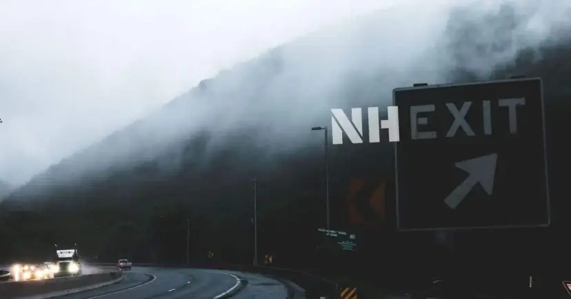 A highway sign reading 'NH Exit' above a foggy mountain road with headlights.