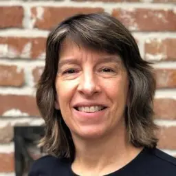 Sheryl Miller, a smiling woman with shoulder-length brown hair, in front of a brick wall.