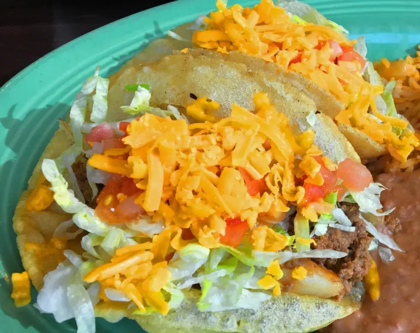 Crispy tacos topped with lettuce, tomato and shredded cheese on a turquoise plate beside refried beans.