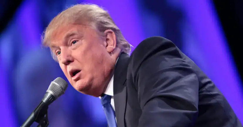 Donald Trump speaking into a microphone against a purple stage backdrop.