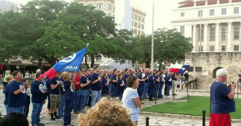 A crowd in navy TNM shirts gathers on Alamo Plaza with blue Alamo Guard flags raised.