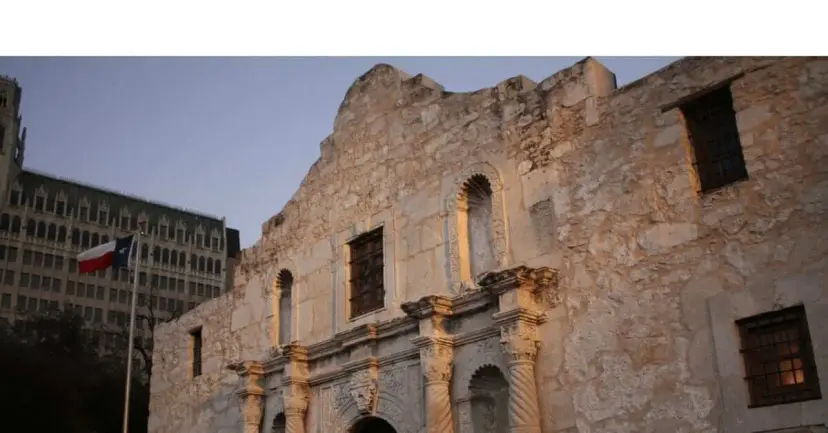 The Alamo church facade at dusk with a Texas flag and a tall hotel building behind it.