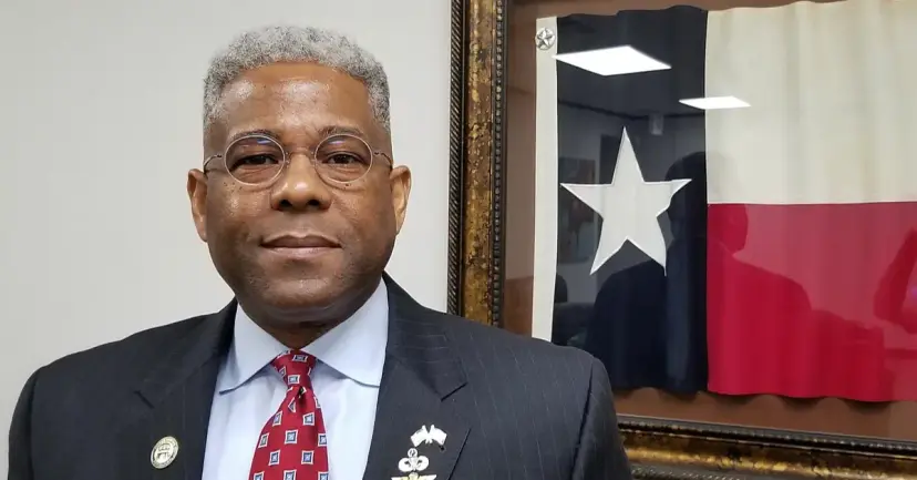 A man in round glasses and a pinstripe suit standing beside a framed Texas flag in an office.