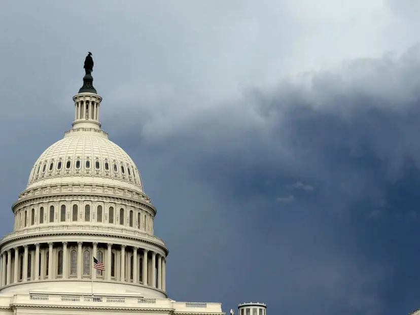 The U.S. Capitol dome in Washington under dark storm clouds, with an American flag flying below.