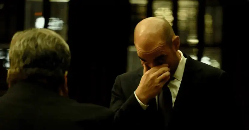 A bald man in a suit covers his face with his hand as another man looks on.