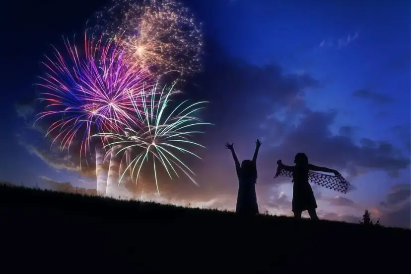 Silhouettes of two children dancing on a hillside at dusk under bursts of colorful fireworks.
