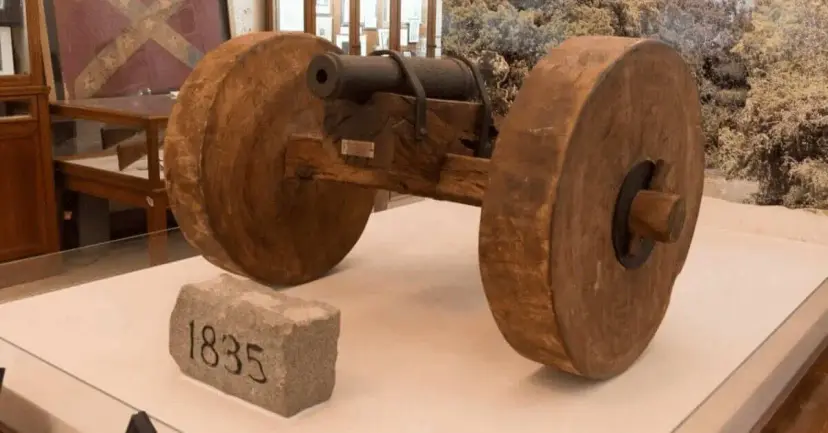 A small historic cannon on wooden wheels displayed in a museum beside a stone marked 1835.