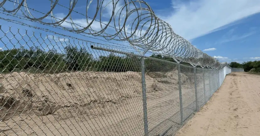A chain link fence topped with coiled razor wire along a sandy border road.