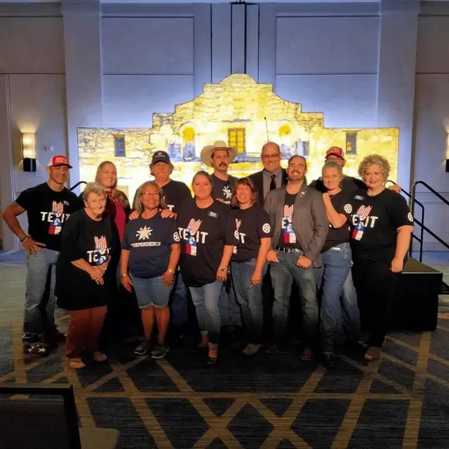 A group in TEXIT and independence shirts poses in front of an Alamo backdrop.