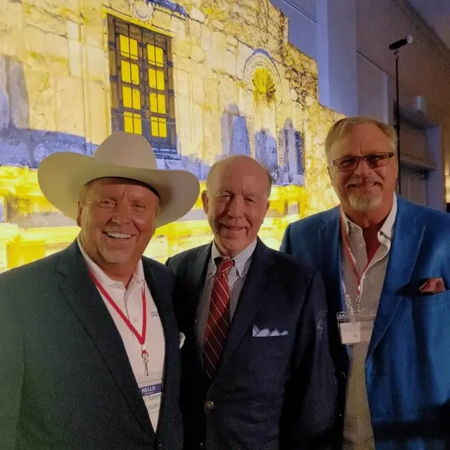 Three men in suits, one in a white cowboy hat, pose in front of an Alamo backdrop.