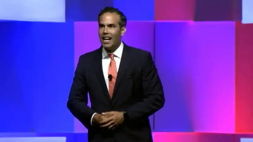 George P. Bush in a dark suit and orange tie speaking on a stage lit in blue and pink.