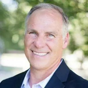 Headshot of Kyle Biedermann smiling outdoors in a dark suit jacket and open collar shirt.