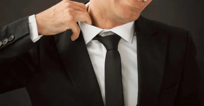 A man in a black suit adjusts his shirt collar and tie.