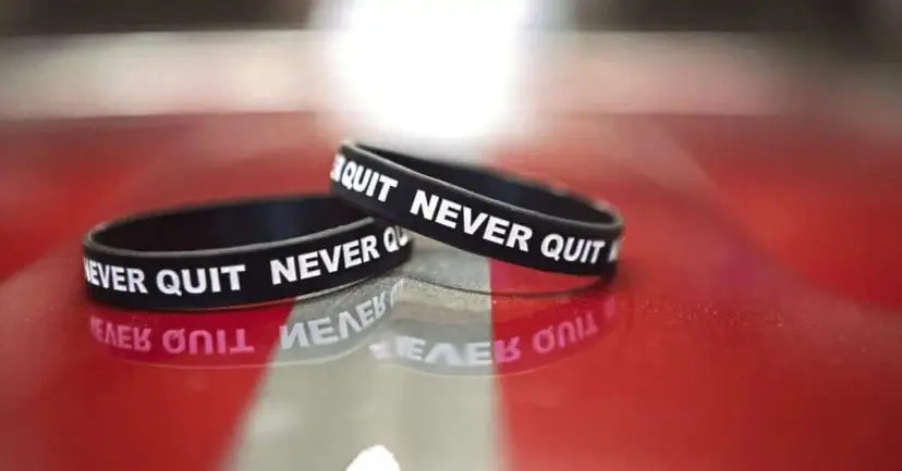 Two black rubber wristbands printed with "Never Quit" resting on a glossy red surface under bright light.
