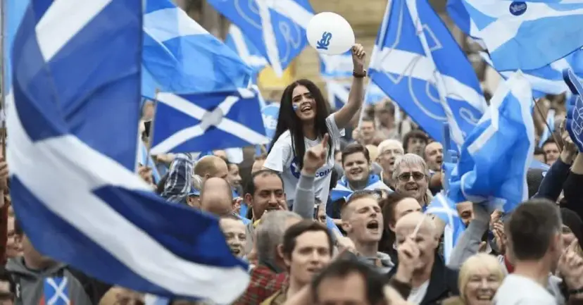 A cheering crowd waves Scottish Saltire flags at a rally as a young woman raises a white balloon.