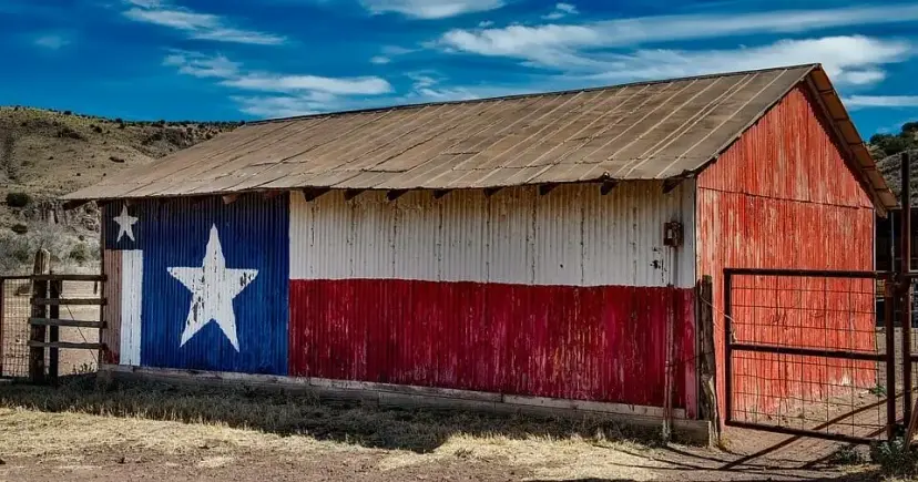 Weathered ranch barn painted with a large Texas flag, set in dry, hilly country under a blue sky.