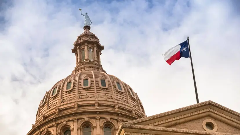 Texas Capitol dome with the Texas flag flying beside it against a cloudy sky.