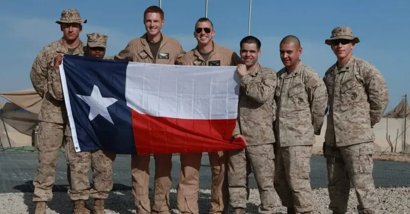 Seven Marines in desert camouflage holding a large Texas flag at a desert military base.
